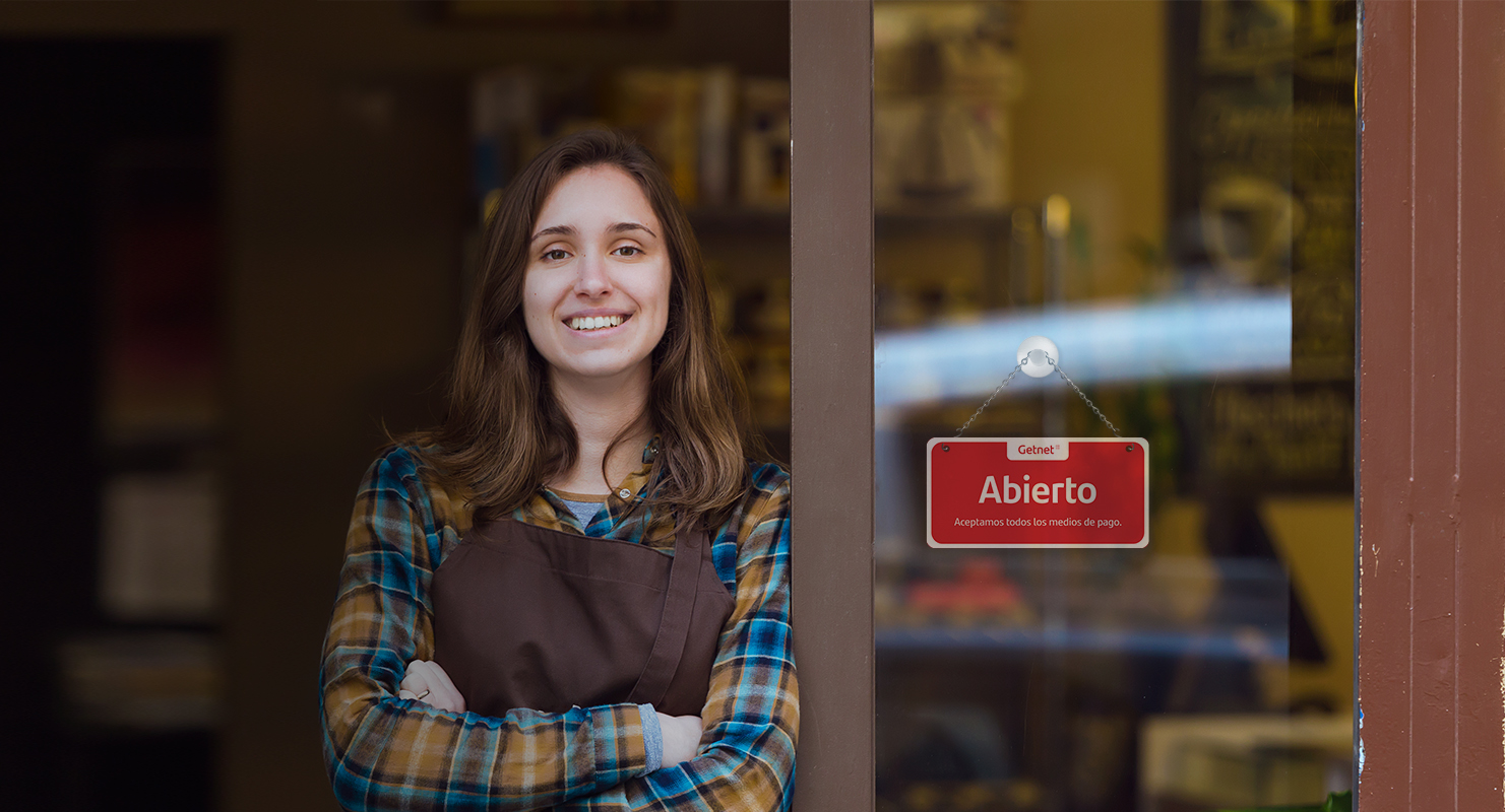 Persona con delantal de pie en la entrada de un local, junto a una puerta de vidrio con cartel rojo de Getnet que dice “Abierto”.