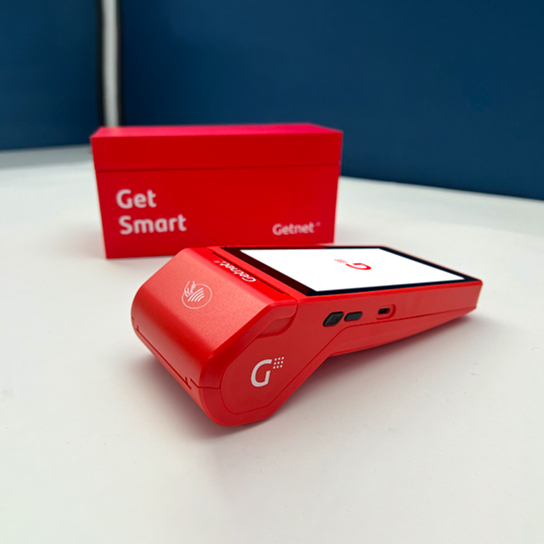 Terminal de cobro Getnet roja apoyada sobre superficie blanca, con pantalla encendida mostrando el logo y caja roja detrás con texto 'Get Smart'.