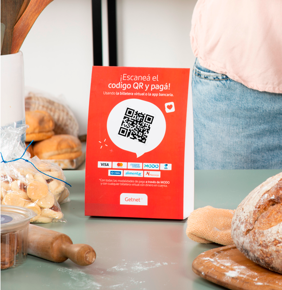 Cartel rojo de Getnet con código QR y texto que indica '¡Escaneá el código QR y pagá!' junto a logos de medios de pago como Visa, Mastercard y MODO, sobre una mesa con panes y utensilios de panadería