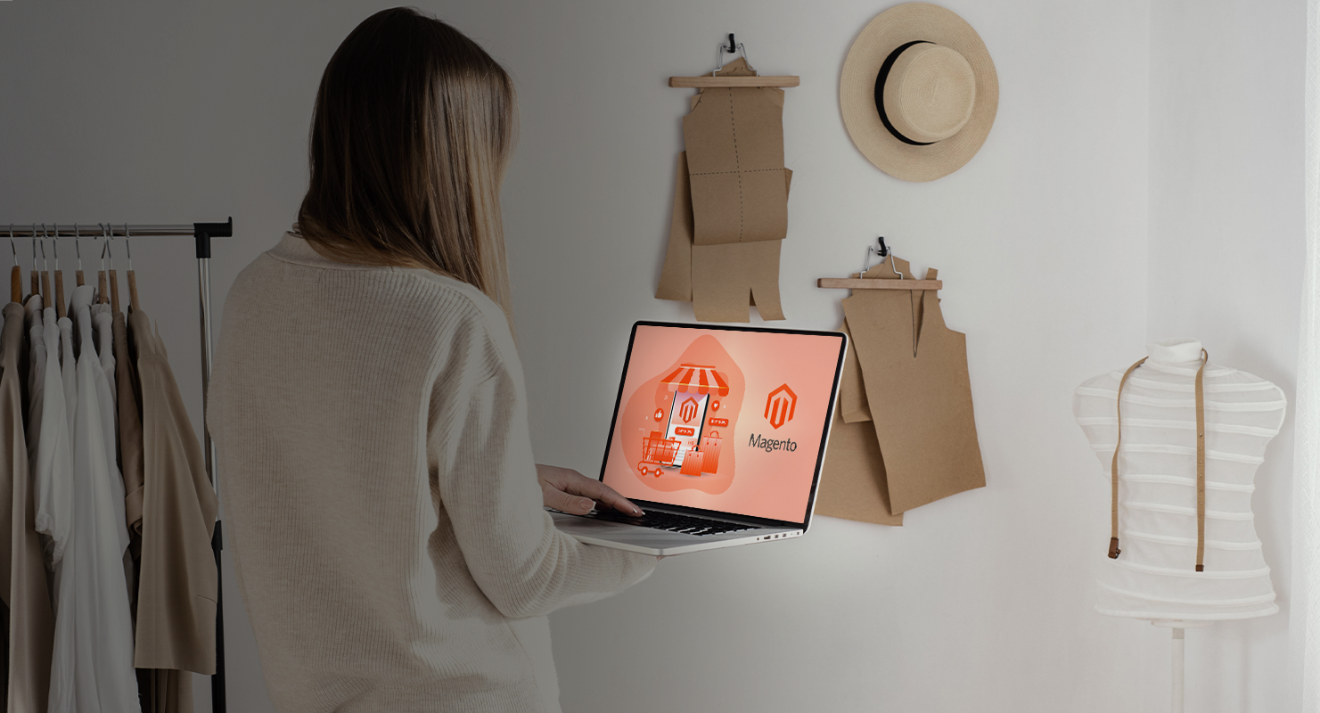 Persona de pie sosteniendo una laptop que muestra el logo de Magento, en una habitación con prendas colgadas, sombrero en la pared y un maniquí.
