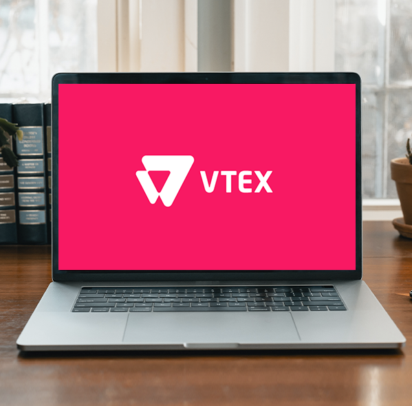 Laptop sobre un escritorio mostrando en la pantalla el logo de VTEX sobre fondo rosa, con libros detrás y una maceta al costado.