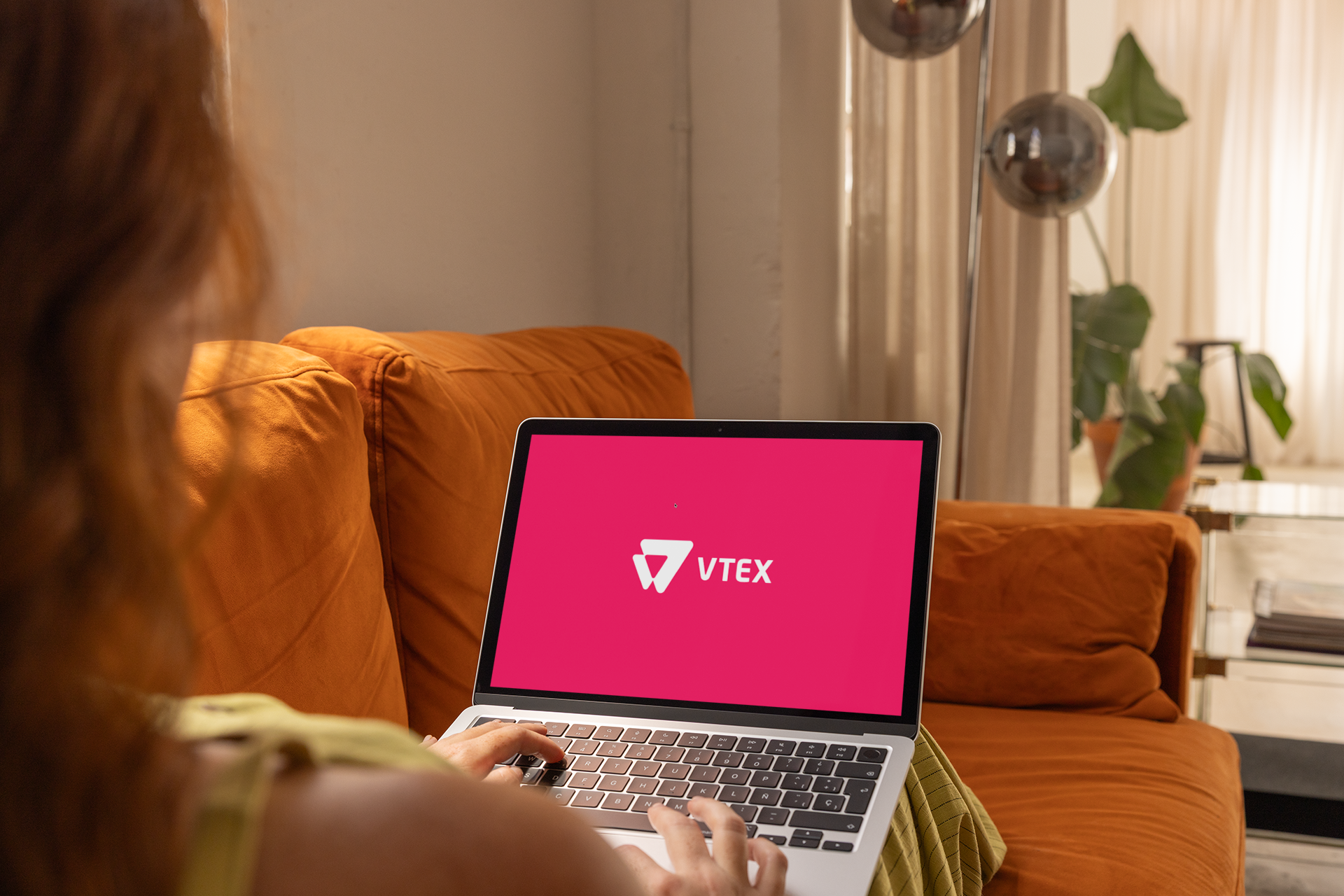 Persona usando una notebook con el logo de VTEX en pantalla, sentada en un sofá naranja.
