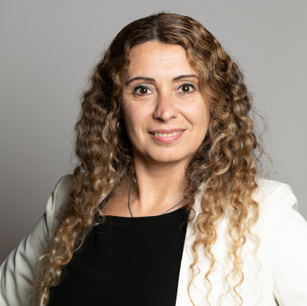 Persona con cabello largo y rizado, usando saco blanco sobre prenda negra, posando frente a un fondo gris.