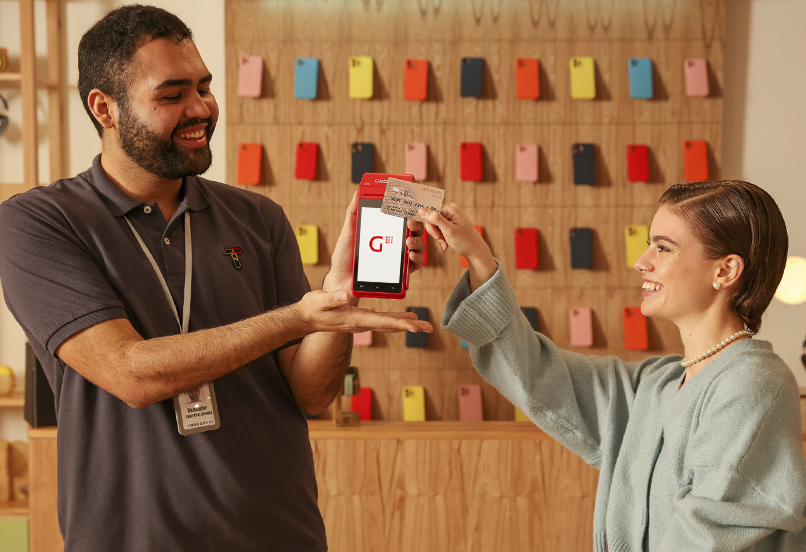 Dos personas en una tienda: una sostiene una terminal GetPOS con el logo de Getnet en la pantalla y la otra apoya una tarjeta en la terminal. Al fondo se ven fundas de celular colgadas en la pared.