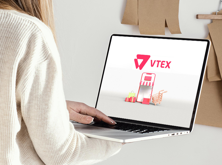 Persona usando una laptop que muestra la pantalla con el logo de VTEX y una ilustración de tienda online con carrito y bolsas de compra.
